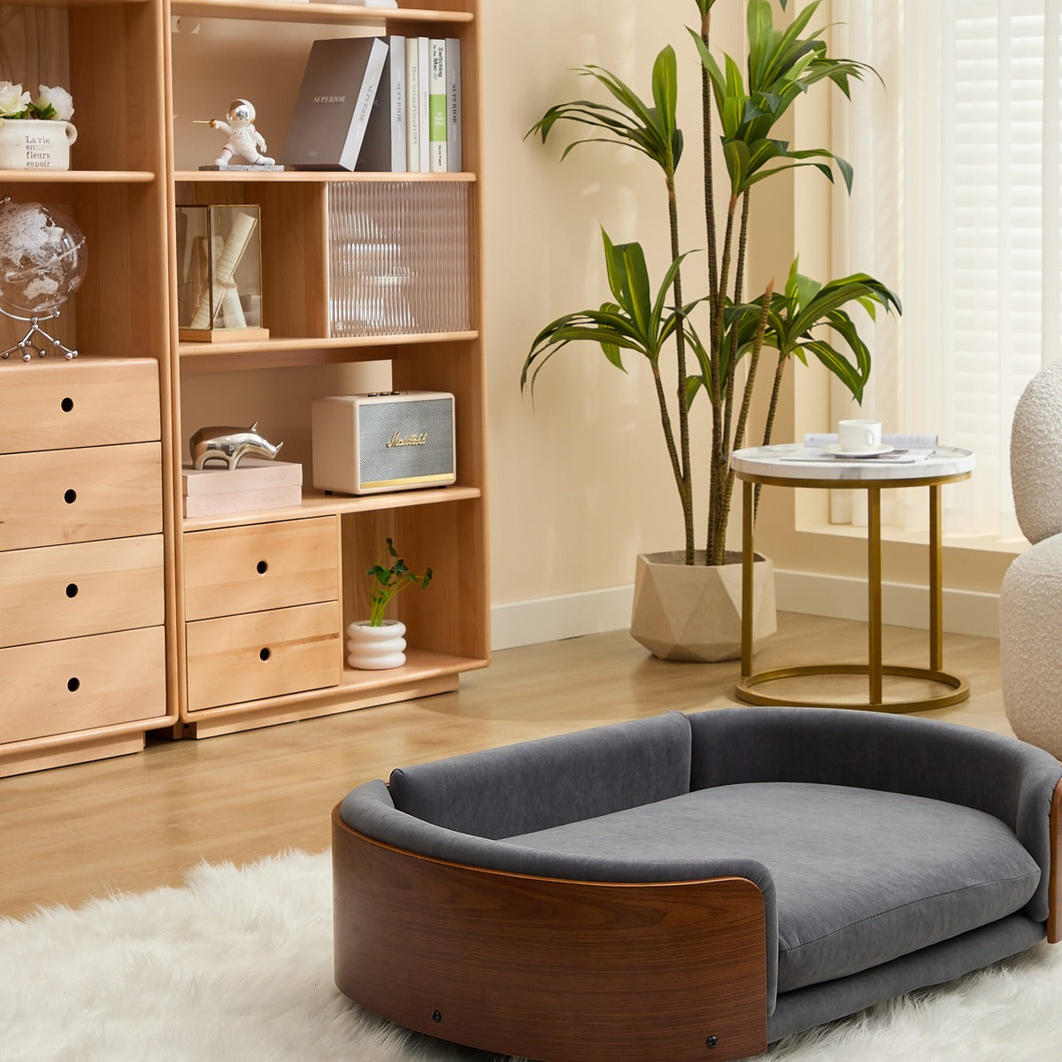 A modern pet bed with a gray velvet cushion and a curved dark wood frame, placed on a white faux fur rug in a bright, contemporary room with a wooden bookshelf, potted plant, and side table.
