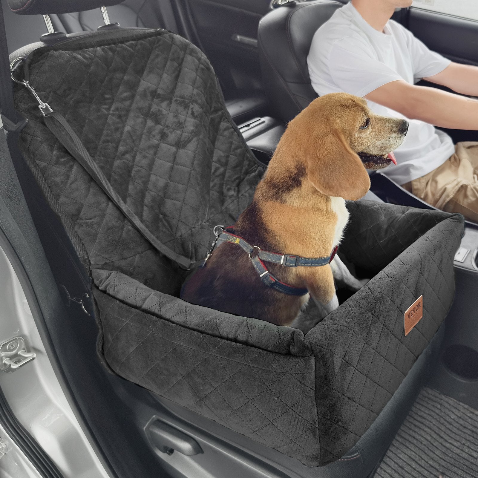 Dog sitting in a black pet seat in a car with a person in the driver's seat.